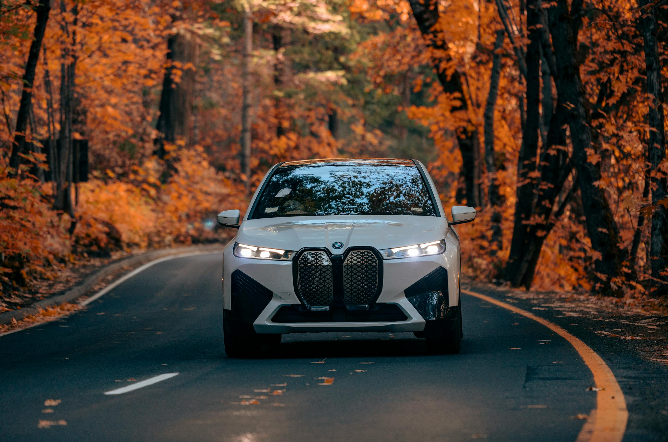 A white car drives through a scenic forest road, surrounded by vivid autumn foliage.