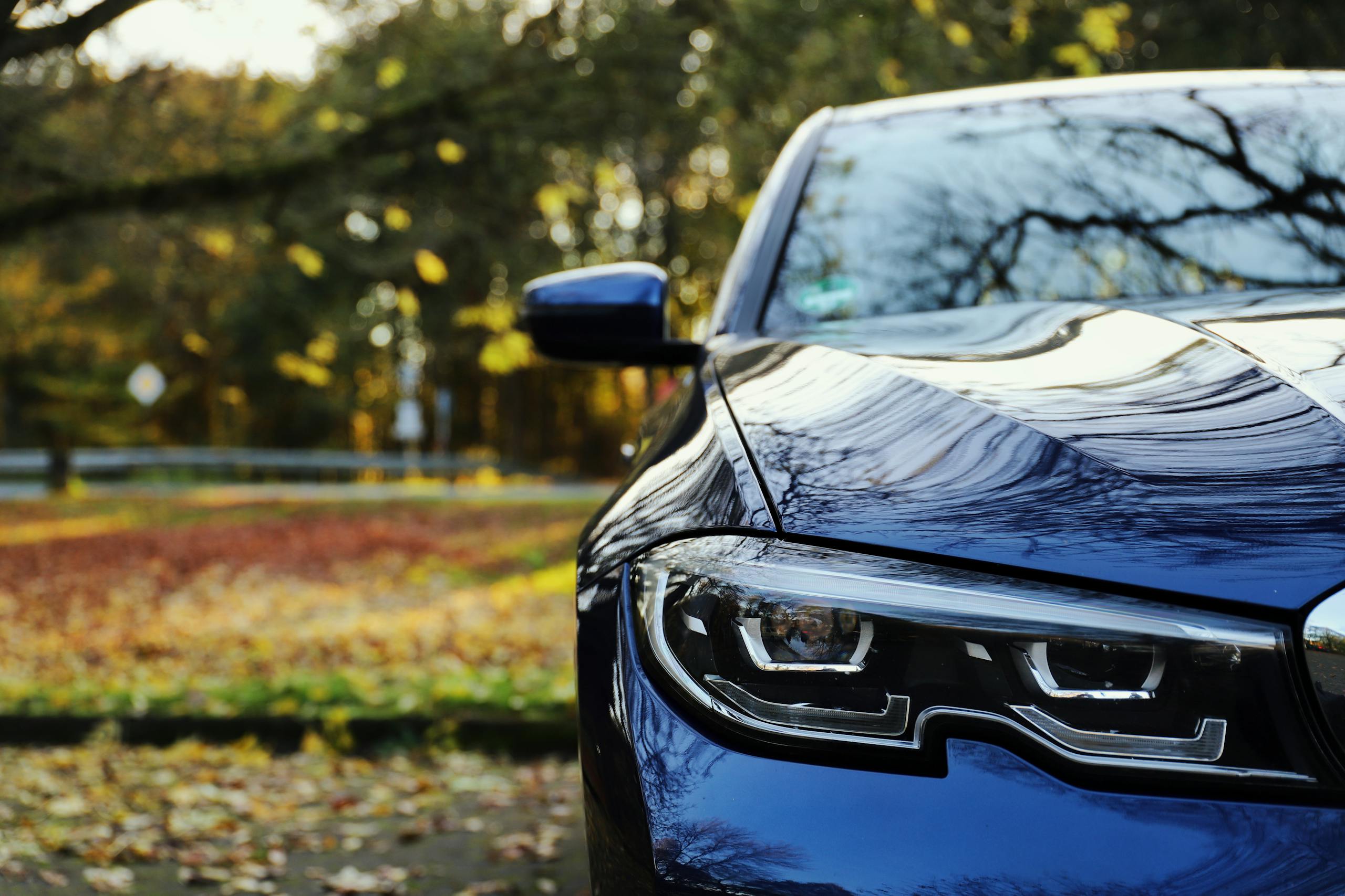 Close-up of a blue car in a parking lot with autumn leaves and reflections on the bonnet.